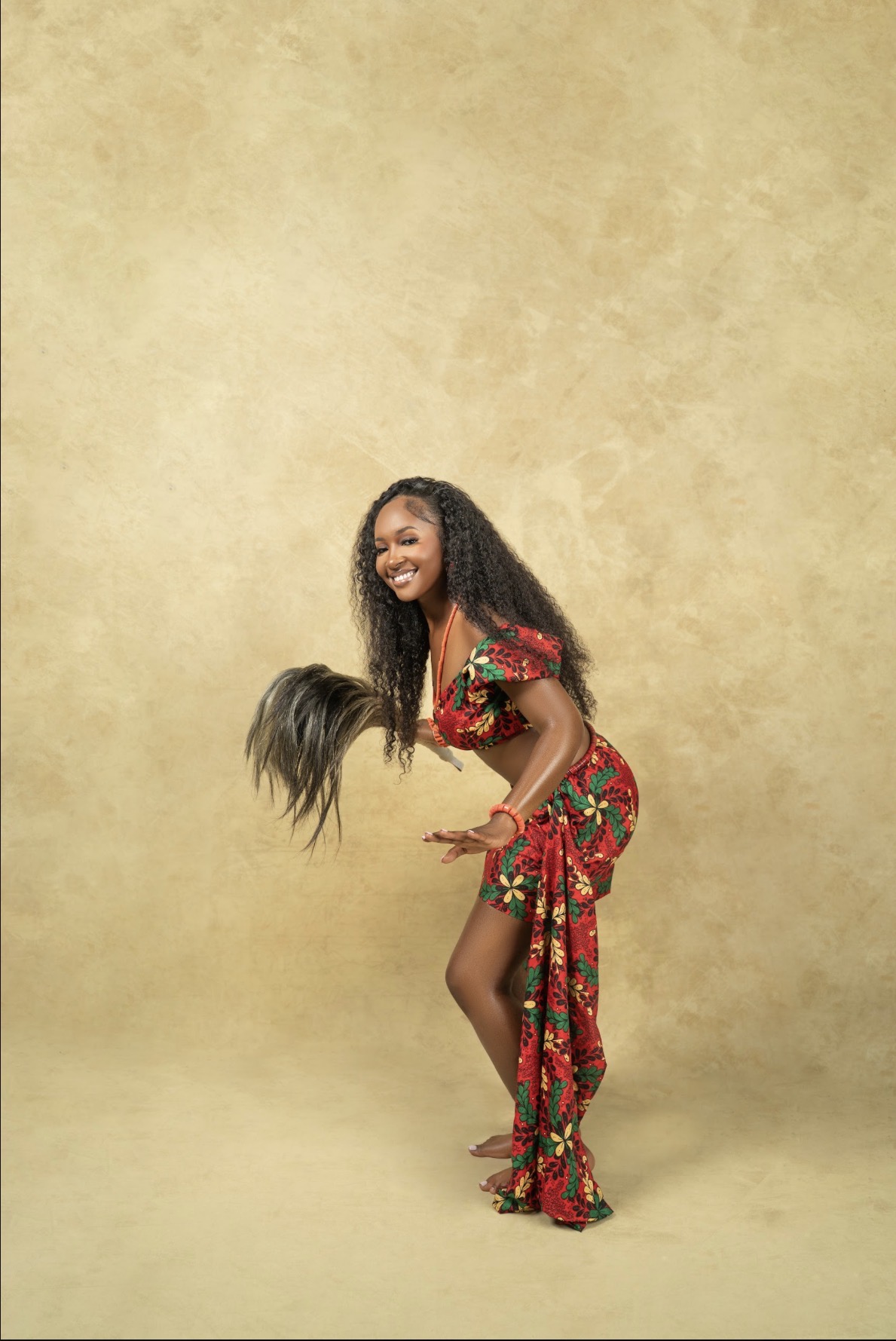Precious dancing in traditional red and green attire with a horse-tail whisk against a warm gold backdrop